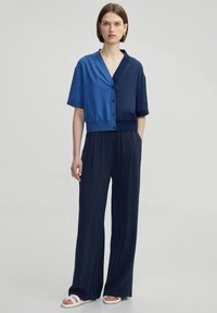Kurzes, ärmelloses Oberteil aus blauem und marineblauem Satin mit Knöpfen. Kombiniert mit weiten, strukturierten, dunkelblauen Hosen und weißen Sandalen.