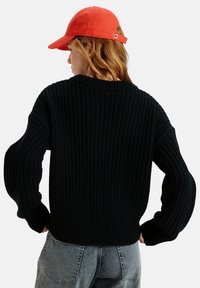 Black knitted sweater with a ribbed texture and dropped shoulder design, complemented by a red adjustable baseball cap.