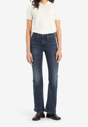 Femme portant un polo blanc à manches courtes, un jean bootcut taille haute bleu foncé et des bottines noires, debout les bras détendus.