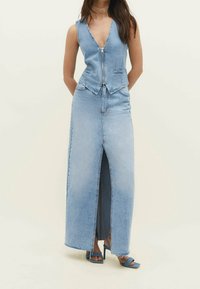 Modelka pózuje ve fitted džískovém vestě a maxi sukni s vysokým rozparkem, doplněná modrými podpatky, na pozadí s jednobarevným světlým pozadím.