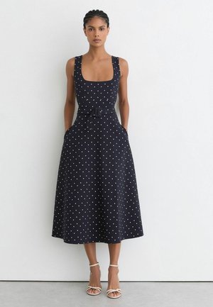 Femme portant une robe midi noire sans manches à pois blancs, taille ceinturée et sandales blanches à talons, debout devant un fond blanc uni.