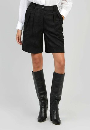Shorts noirs à rayures avec des plis, associés à des bottes en cuir noires à talons hauts, arrivant jusqu'aux genoux. Le tissu est lisse avec une coupe structurée.