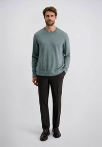 Helles grün gestricktes Sweater mit rundem Ausschnitt, kombiniert mit dunkelbraunen maßgeschneiderten Hosen und dunklen Slippern, vor einem neutralen Hintergrund.