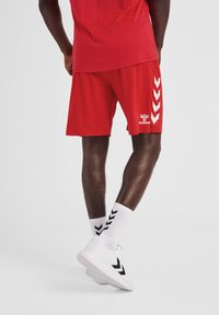 Pantalones cortos deportivos rojos con un corte relajado, que presentan un logo blanco y un patrón en forma de Chevron en el lateral. Combinados con calcetines blancos y zapatillas.