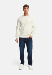 Maglione bianco a trame con strisce orizzontali, abbinato a jeans blu scuro e sneakers bianche, esposto su una figura maschile in piedi.