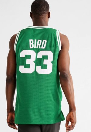 Hombre con una camiseta de baloncesto verde con ribete blanco, número 33 y el nombre Bird en la espalda, de pie frente a un fondo blanco liso.