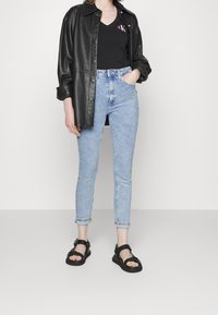 Chaqueta de camisa de cuero negra, camiseta de cuello en v negra con logotipo, jeans ajustados de longitud tobillo en azul claro, sandalias planas negras con tiras.