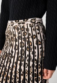 Vrouw in een rokje met luipaardprint en plooien met verticale strepen, gecombineerd met een zwart kabelgebreid sweater, hand relaxed langs de zijde.