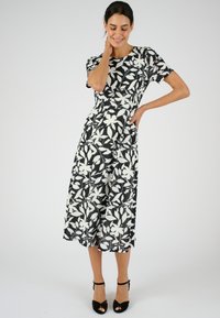 Vestido con estampado floral que presenta hojas en blanco y negro, mangas cortas, un corpiño ajustado y una falda larga y fluida de longitud media. Sandalias de tacón negro.