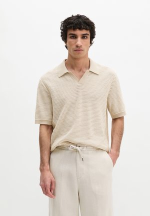 Man with curly dark hair wearing a beige knit short-sleeve polo and light beige drawstring pants, standing against a plain white background.