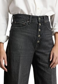 Jeans de mezclilla negros de cintura alta con cierre de botón frontal, llevados con una camisa blanca de manga larga metida dentro del pantalón, con las manos relajadas a los lados.