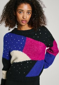 Mehrfarbiger Strickpullover mit schwarzen, pinken, blauen und cremefarbenen Segmenten, verziert mit silbernen Pailletten auf einem strukturierten Stoff.