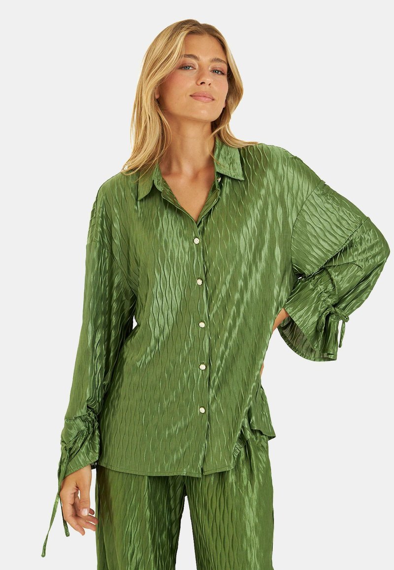 Camisa de satén verde con un patrón texturizado y ondulado, mangas largas, cierre de botones en el frente y puños ajustables con cordón. La tela tiene un acabado brillante.