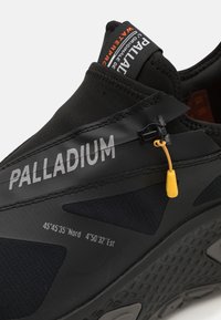 Mustad spordijalats, millel on veekindel ülemine osa, tekstuuriga paneelid, kollased aktsendid ja silmapaistvalt paigutatud "PALLADIUM" logo küljepeal.