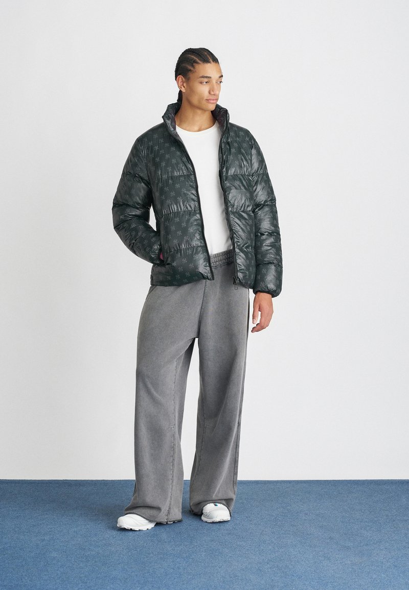 Grüne Steppjacke mit glänzender Oberfläche und dezenter Musterung, ausgestattet mit einem hohen Kragen und einem Reißverschluss vorne, kombiniert mit grauen weit geschnittenen Hosen.