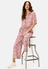 Bloemen jumpsuit in tinten van roze, oranje en wit. Korte mouwen, knoopsluiting aan de voorkant, elastische tailleband, wijde broekspijpen, gecombineerd met witte sneakers.