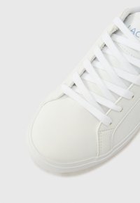 Λευκό αθλητικό παπούτσι με λείο δέρμα, στρογγυλεμένη μύτη, επίπεδα κορδόνια και διακριτικό ραφτό σχέδιο στο πλάι. Απλός σχεδιασμός με ελάχιστες διακοσμήσεις.