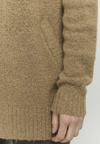 Beige, texturerad stickad tröja med ribbad mudd och nederkant, med en sidofickdetalj. Tyget verkar mjukt och varmt.