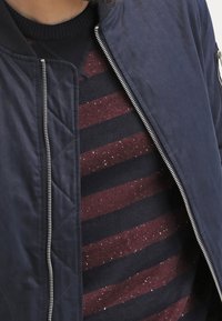 Mann trägt eine marineblaue gesteppte Jacke, offen über einem burgunderroten und marineblau gestreiften Pullover mit weißen Sprenkeln.