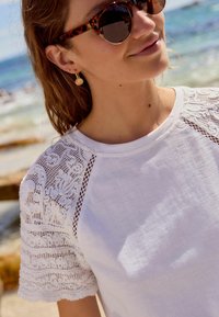 T-shirt bianco con dettagli in pizzo intricati sulle maniche corte. Lo scollo è rotondo e il tessuto appare leggero e liscio.