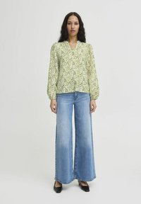Blusa floreale verde con maniche a sbuffo lunghe e scollatura a V, abbinata a jeans azzurri a zampa d'elefante. Scarpe nere a punta completano l'outfit.