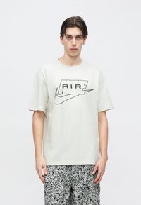 Tânăr purtând un tricou alb cu logo-ul Nike Air și pantaloni scurți cu model alb-negru, stând pe un fundal alb simplu.