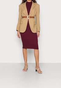 Blazer beige con spalline, indossato sopra un abito aderente bordeaux, cintrato in vita con una cintura marrone e fibbia argentata. Sandali beige con cinturini.