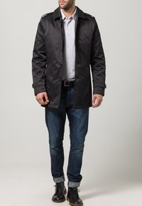 Svart vattentät trenchcoat med knappstängning, axelremmar och fodrad insida, ihop med denimjeans och svarta kängor.