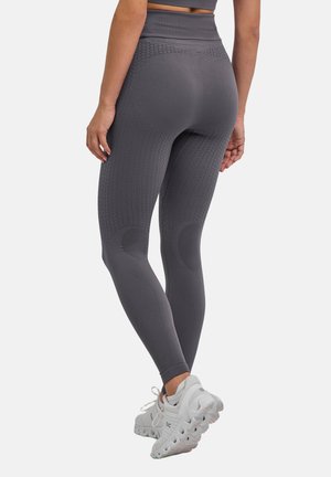 Frau trägt graue, hoch taillierte Leggings mit Struktur und weiße Laufschuhe, von hinten vor einfachem Hintergrund gezeigt.