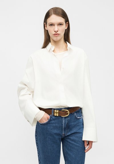 Young woman with straight brown hair wears oversized white shirt, blue jeans, and brown belt with gold buckle, standing against plain background.