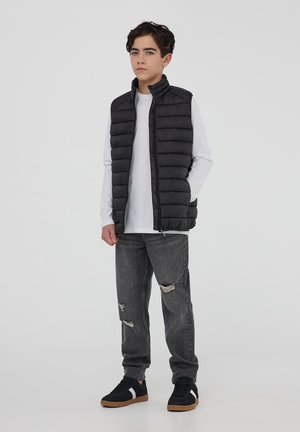 Czarna pikowana kamizelka z wysokim kołnierzem, noszona na białej koszuli z długim rękawem. Dobrana do podniszczonych szarych dżinsów i czarnych sneakersów z gumową podeszwą.