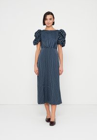 Robe bleu marine à pois avec manches bouffantes, taille ajustée et jupe mi-longue. Texture lisse et motif de pois blancs distincts.