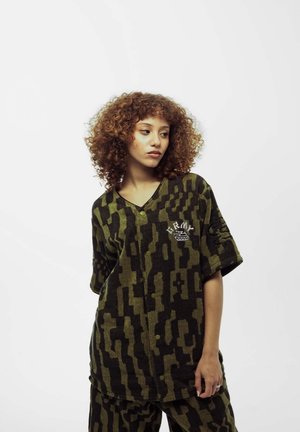 Camisa de gran tamaño con patrón de camuflaje en verde olivo y negro, con cierre de botones en la parte anterior y detalle de logotipo en el pecho. Textura suave.