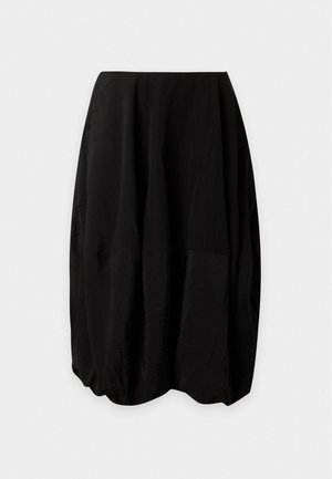 Femme debout portant un chemisier blanc sans manches à boutons, une jupe midi noire volumineuse et des ballerines noires sur fond blanc uni.