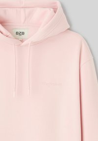 Sweat à capuche rose doux avec cordon de serrage, poignets côtelés, et le mot "Perfezione" délicatement brodé sur le devant. Design décontracté et confortable.