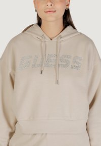 Felpa beige con cappuccio caratterizzata da un fit morbido e orlo a costine, con la scritta "GUESS" in strass argentati sul petto.