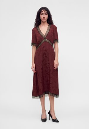 Femme aux cheveux bouclés portant une robe midi bordeaux avec bordure en dentelle noire et des escarpins noirs, debout devant un fond blanc uni.