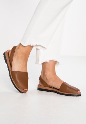 Sandales à talon ouvert en cuir marron portées avec un pantalon blanc à ourlet effiloché sur un sol blanc lisse.