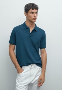 Mladý muž ve tyrkysovém polo tričku s krátkým rukávem a bílých kalhotách, stojící s jednou rukou v kapse, dívající se na stranu proti jedné holé zdi.