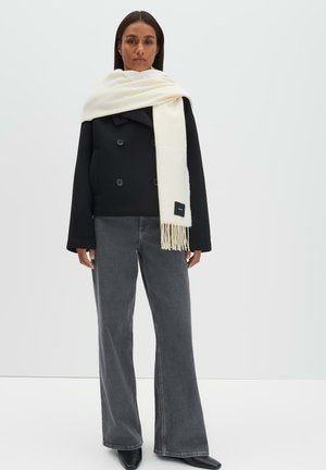 Manteau noir croisé à double boutonnage, écharpe blanche drapée sur les épaules, jean gris à jambes larges, et chaussures noires pointues, debout devant un fond blanc.