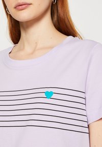 Femme portant une chemise violet clair avec des rayures horizontales noires sur la poitrine et un petit cœur bleu centré sur les rayures.