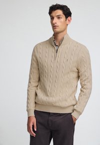 Beige stickad tröja med hög krage, med ett flätmönster. Ribbad ärm- och nederkant, med dragkedja och diskret logotypdetalj.