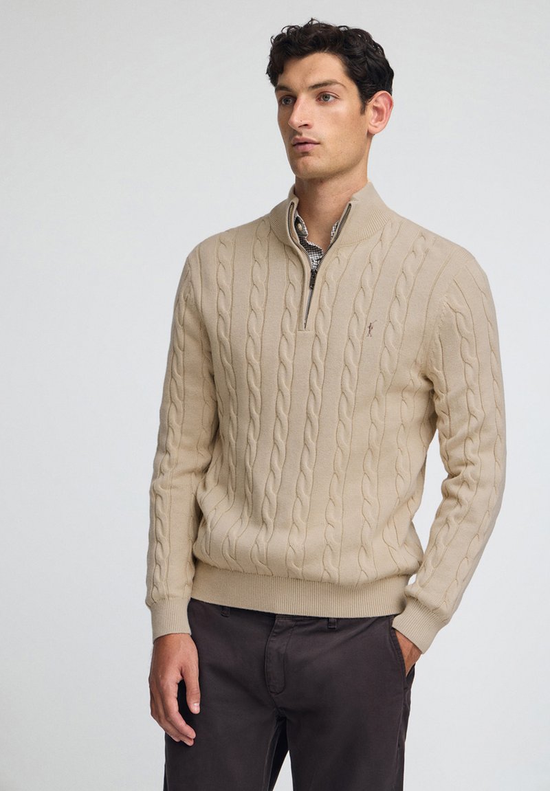 Beige stickad tröja med hög krage, med ett flätmönster. Ribbad ärm- och nederkant, med dragkedja och diskret logotypdetalj.