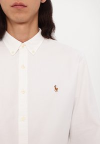 Buste vêtu d'une personne portant une chemise blanche boutonnée avec un petit logo brodé représentant un cheval et un cavalier sur le côté gauche de la poitrine.