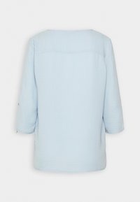 Blusa azzurro chiaro, con texture, dal collo rotondo e maniche a tre quarti. Presenta una cucitura a yoke sul retro e dettagli con bottoni sulle maniche.