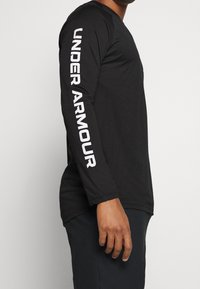 Czarna koszulka z długim rękawem wykonana z mieszanki bawełny i poliestru, z białym logo "UNDER ARMOUR" na lewym rękawie, okrągłym dekoltem i luźnym krojem.