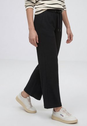 Osoba nosząca czarne spodnie z szerokimi nogawkami ze sznurkiem w pasie i białe trampki, stojąca na tle jednolitego jasnego tła.