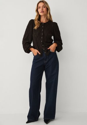 Kvinde med langt blondt hår iført en sort tekstureret knappet bluse med puffede ærmer og mørke brede jeans, står med hænderne i lommerne.