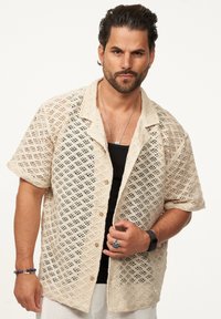 Beige Leinenhemd mit kurzen Ärmeln, das ein diamantförmiges Perforationsmuster aufweist, mit Kragen und Knopfleiste, kombiniert mit einem schwarzen Tanktop.
