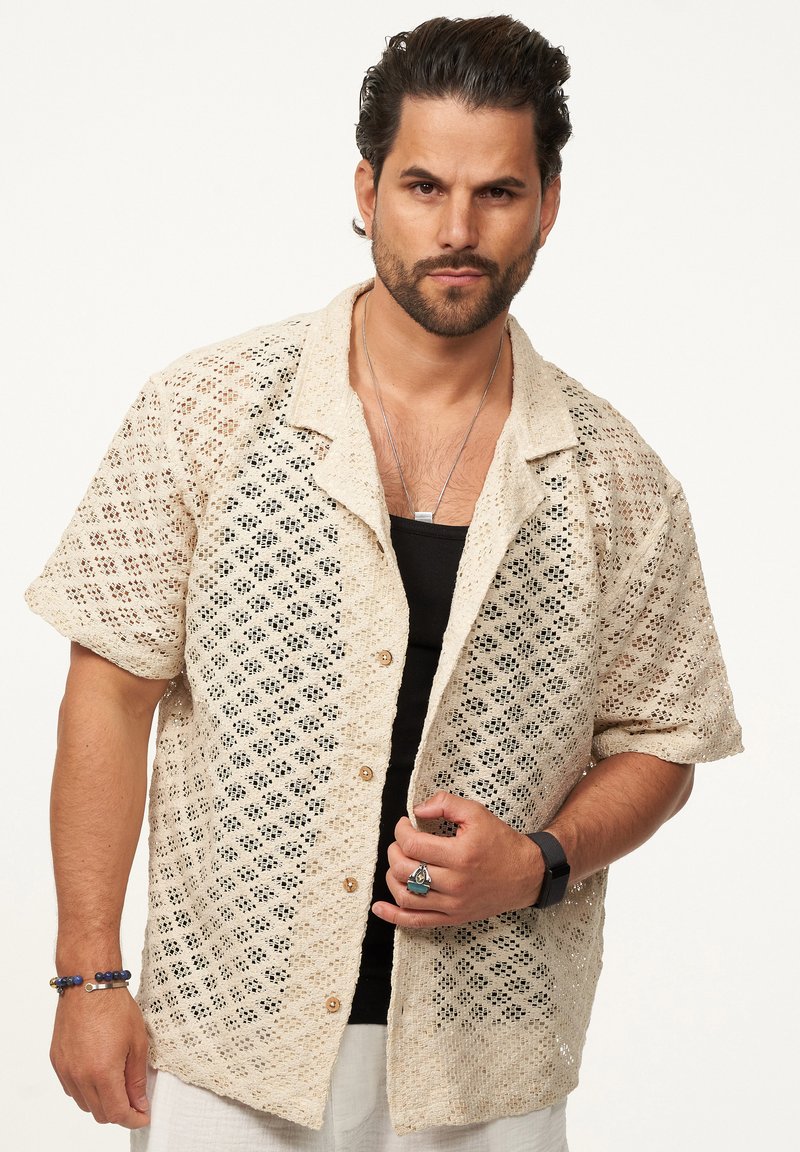 Beige Leinenhemd mit kurzen Ärmeln, das ein diamantförmiges Perforationsmuster aufweist, mit Kragen und Knopfleiste, kombiniert mit einem schwarzen Tanktop.